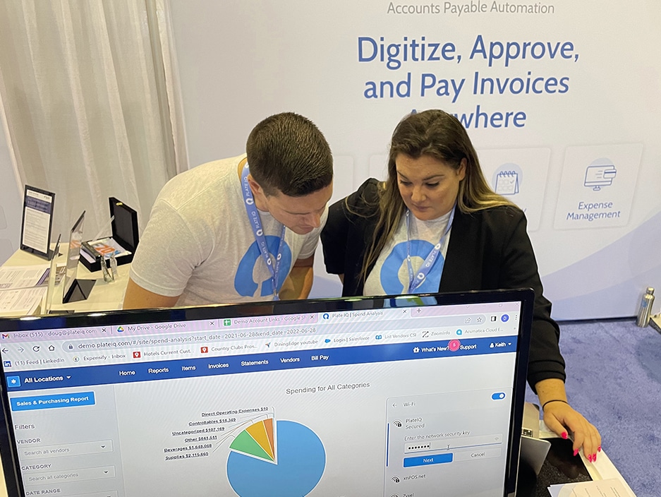 Colleagues adjust a demo screen at Plate IQ's booth in Orlando, Florida, at the HITEC hospitality conference for hotels and country clubs. Background explains that Plate IQ's accounts payable automation means that hotel groups and country clubs can digitize, approve and pay invoices from anywhere. 